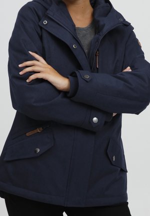 Persoon draagt een marineblauwe capuchonjas met zilveren drukkers, ritssluitingszakken en armen gekruist over een grijs shirt.