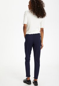 Sieviete ar cirtainiem matiem, tērpusies baltā ribotā īsajās piedurknēs blūzē, tumši zilās uzstumtas biksēs un melnās aukliņu kurpēs, stāvot ar rokām kabatās.