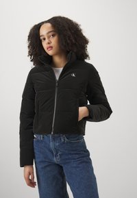 Sort pufferjakke med høj krave og lynlås, der har et syet mønster. Modellen kombinerer den med højtaljede blå jeans.