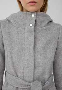 Manteau en mélange de laine gris avec une capuche structurée, comportant deux fermetures à pression métalliques et une ceinture à la taille. Motif en chevron texturé visible.