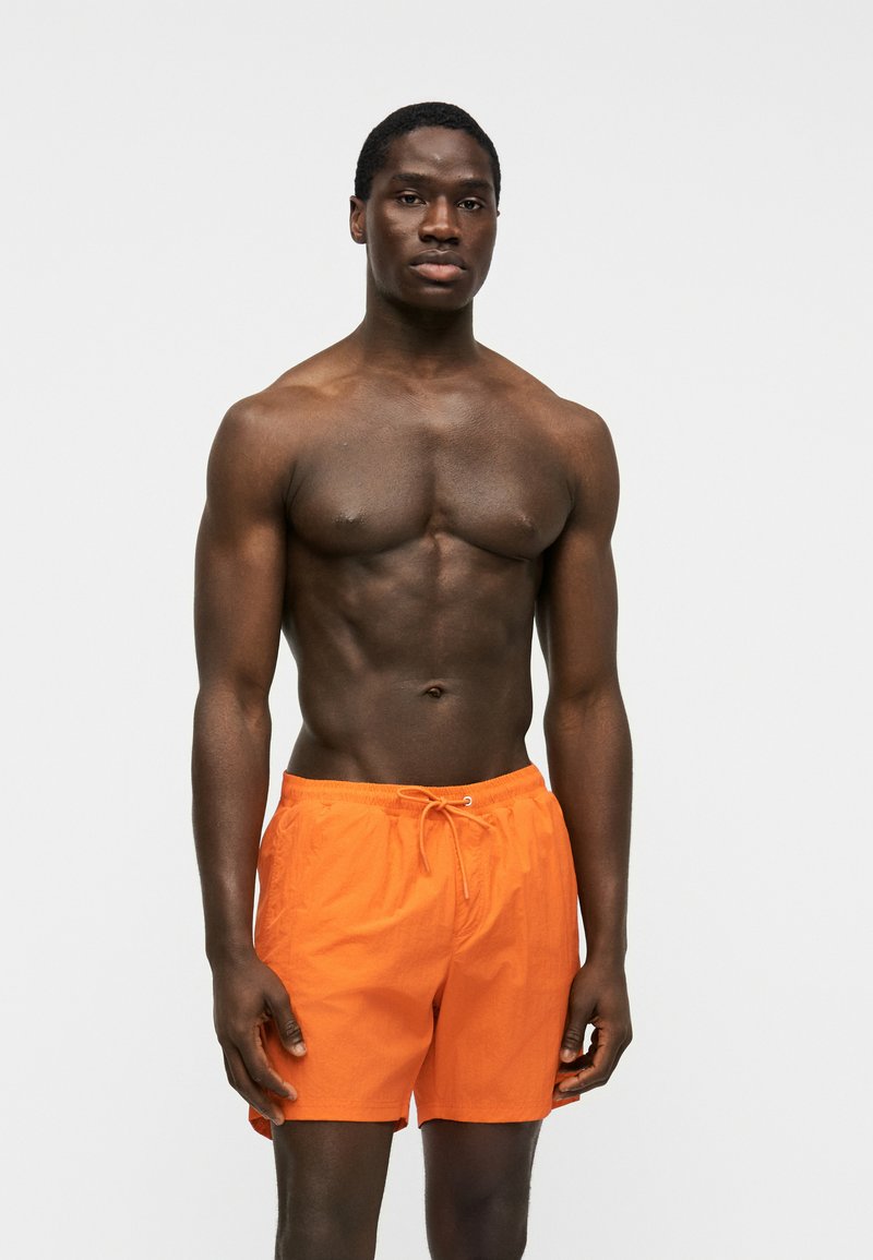 Homme torse nu avec une silhouette musclée portant un short de bain orange vif, debout sur un fond uni gris clair, regardant vers l'avant.