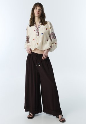 Jeune femme portant un chemisier crème brodé et un pantalon large noir, debout, les mains dans les poches, devant un fond uni.