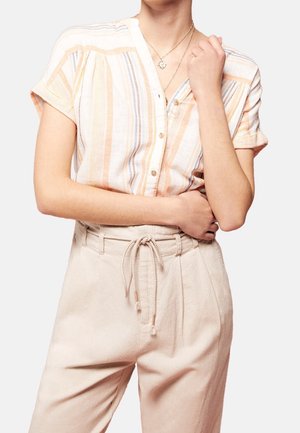 Persoon met een kortmouwig, gestreept overhemd met knopen en beige broek met hoge taille, vastgebonden met een touwriem, staand met gekruiste armen.