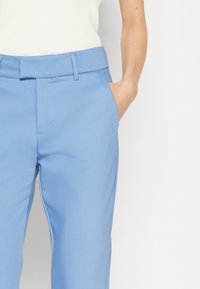 Světle modré kalhoty s hladkým povrchem, předními kapsami a rovný střih. Obsahují poutka na opasek pro nastavitelnou velikost.