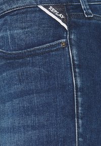 Jeans de denim azul oscuro con un bolsillo frontal, que presentan una etiqueta blanca y azul que dice "REPLAY". Textura suave y ajuste estándar.
