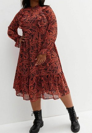 Kvinde iført en orange og sort paisley midi-kjole med lange ærmer, kombineret med sorte snørelærredsstøvler, står op ad en hvid væg.