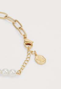 Pulseira de corrente de ouro com contas brancas redondas, um fecho de lagosta e uma etiqueta decorativa com um logótipo gravado. Textura suave em geral.