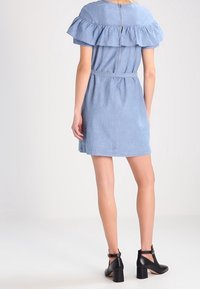 Robe en denim bleu clair avec un design à volants en dehors des épaules, taille cintrée et fermeture éclair au dos. Associée à des bottines noires.