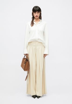 Cremefarbene plissierte Hose kombiniert mit einem lockeren weißen Hemd, akzentuiert durch eine braune Handtasche und schwarze spitze High Heels.