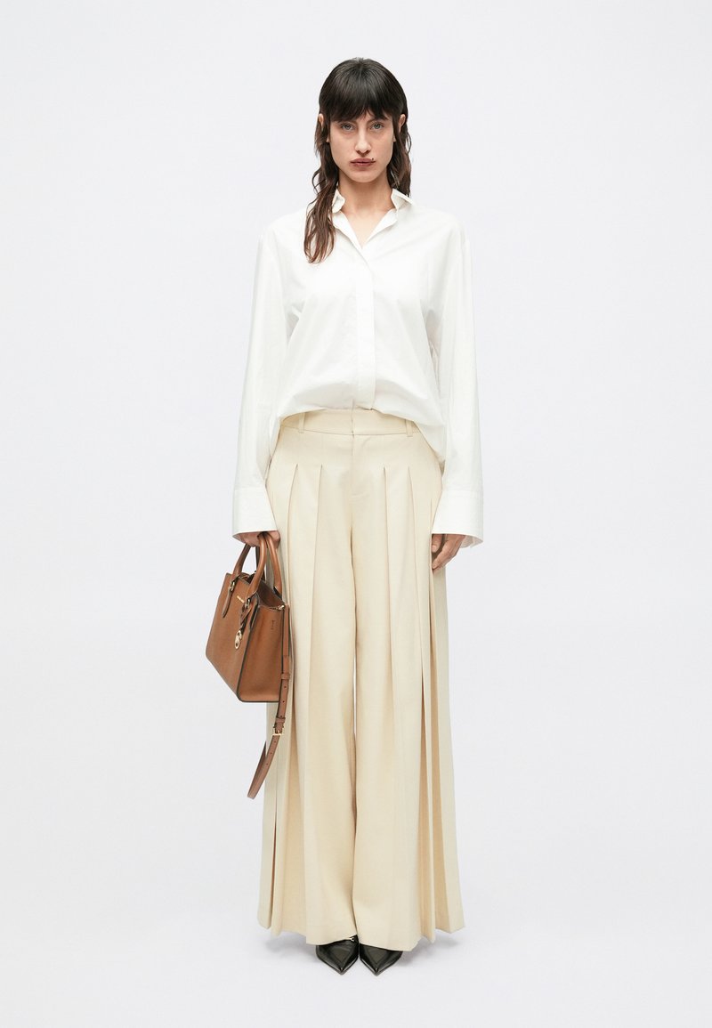 Κρεμ παντελόνι με πιέτες συνδυασμένο με λευκό, άνετο πουκάμισο με κουμπιά, τονισμένο από καφέ τσάντα χειρός και μαύρες μυτερές γόβες.