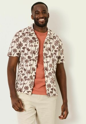 Lächelnder Mann, der ein kurzärmeliges Hemd mit Palmenmuster über einem korallenfarbenen T-Shirt und hellen Hosen trägt.