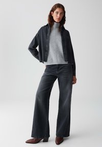 Junge Frau trägt einen grauen Rollkragenpullover, eine schwarze Jeansjacke, weite schwarze Jeans und braune spitze Stiefel und steht vor einem weißen Hintergrund.