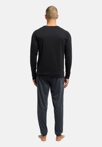 Mand står barfodet med ryggen til, iført en sort langærmet skjorte og mørkegrå sweatpants foran en ensfarvet lys baggrund.