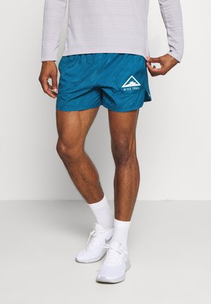 Mężczyzna w niebieskich spodenkach do biegania Nike Trail z wzorem drewnianej faktury, białych skarpetkach, białych butach sportowych Nike i jasnoszarej bluzce z długim rękawem.