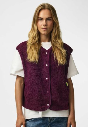 Jeune personne aux longs cheveux blonds portant une chemise blanche et un gilet polaire violet boutonné, debout devant un fond blanc uni.