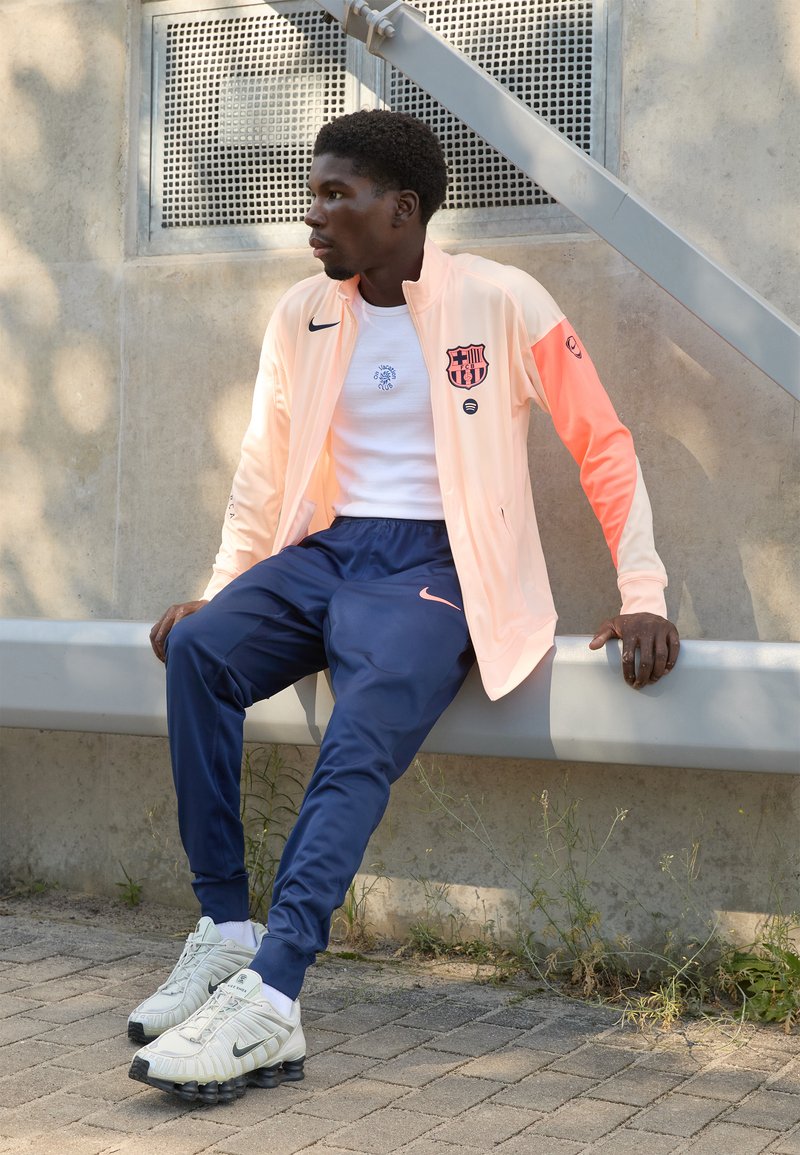 Light peach sports jacket with a logo, white shirt, navy pants, and white shoes with black accents. Athlete seated on a bench.