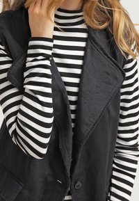 Gilet sans manches noir avec un col châle et des bords effilochés, porté sur un haut à manches longues noir et blanc rayé. Détail de bouton visible sur le gilet.