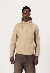 Beige huvtröja med dragkedja framtill, justerbara dragsnören och elastiska ärmslut. Har en liten vit logotyp på vänster bröst.