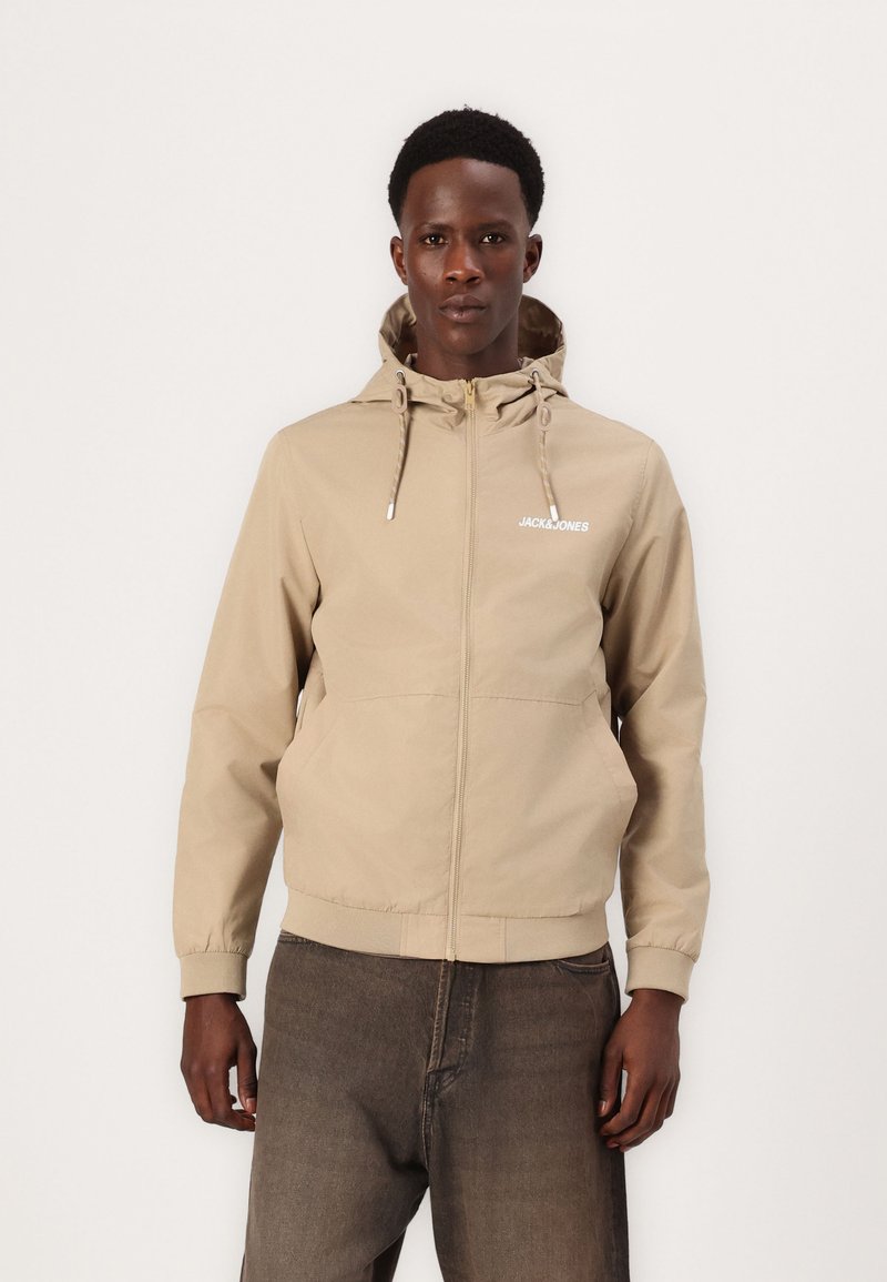 Beige huvtröja med dragkedja framtill, justerbara dragsnören och elastiska ärmslut. Har en liten vit logotyp på vänster bröst.