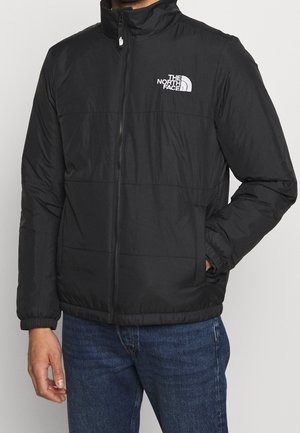 Homme portant une veste zippée noire North Face, les mains partiellement dans les poches, et un jean bleu sur un fond clair uni.