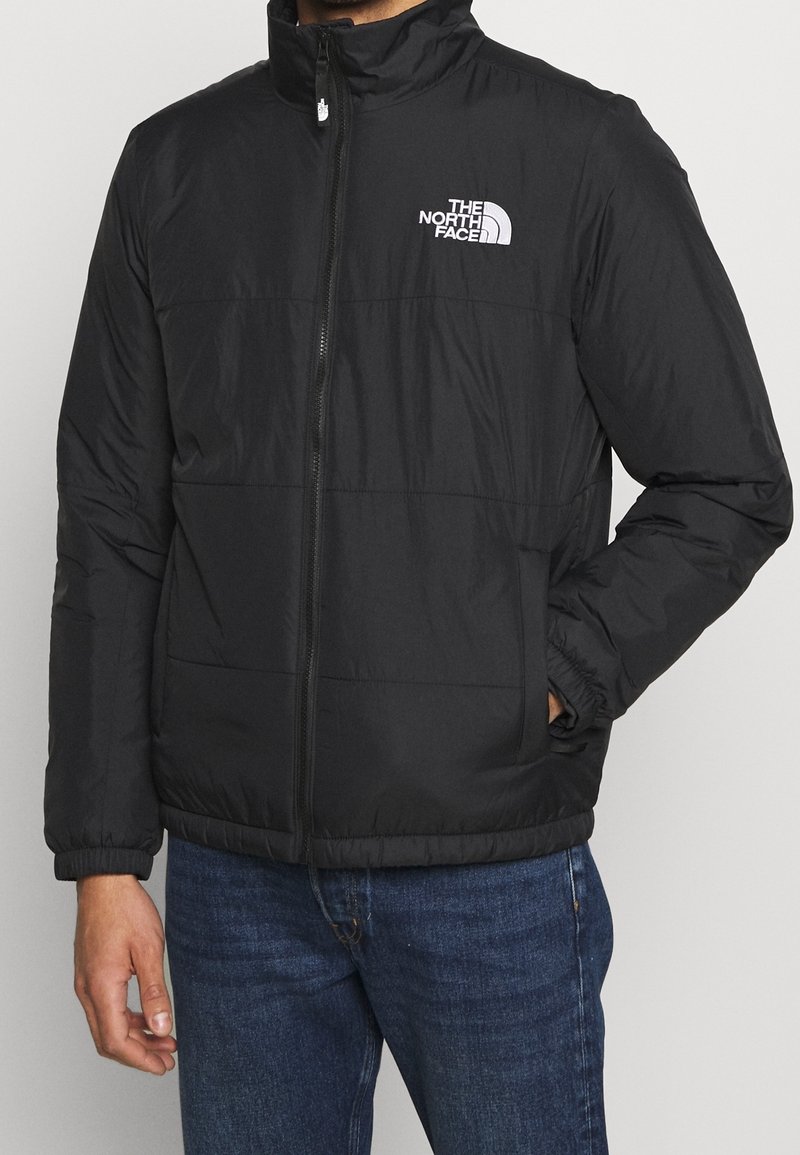 Homme portant une veste zippée noire North Face, les mains partiellement dans les poches, et un jean bleu sur un fond clair uni.