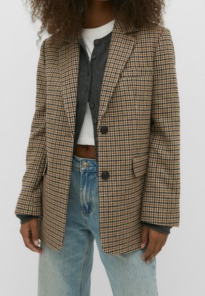 Frau trägt einen braun-beigen karierten Blazer über einem dunkelgrauen, geknöpften Cardigan und einem weißen, bauchfreien Top, kombiniert mit hellblauen Jeans.
