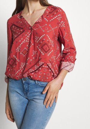 Vrouw draagt een rood patroon langemouwen blouse met V-hals en lichtblauwe spijkerbroek, één hand rustend op haar heup.