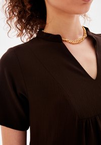 Braunes, strukturiertes Kleid mit V-Ausschnitt und kurzen Ärmeln. Eine goldene Kettenhalskette fügt einen metallischen Akzent am Ausschnitt hinzu.
