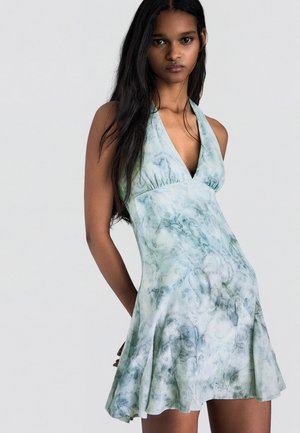 Femeie cu păr lung și închis la culoare, purtând o rochie fără mâneci, bleu-verzui deschis, cu un model floral subtil, stând pe un fundal simplu.