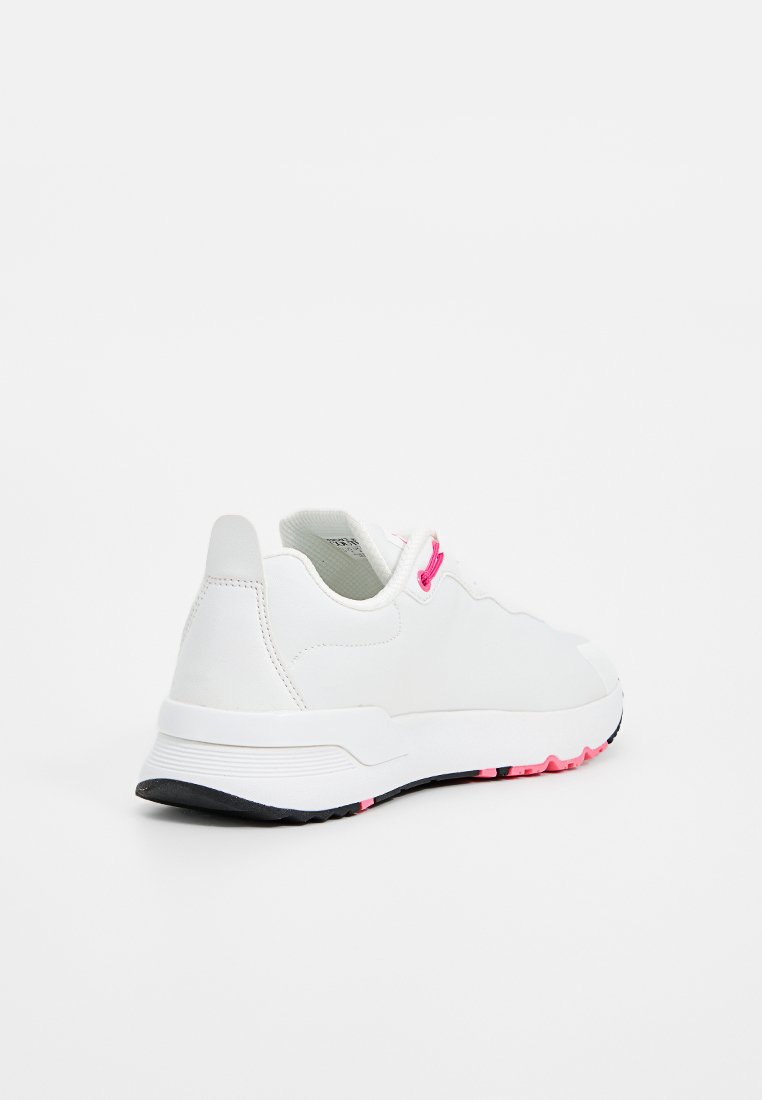 Zapatilla deportiva blanca con lazos de color rosa y suela de tracción negra y rosa, mostrada desde el lado posterior izquierdo sobre un fondo blanco.