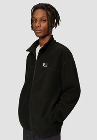 Schwarze Fleecejacke mit hohem Kragen, Reißverschluss vorne und Logopatch auf der Brust. Weicher Stoff und lockere Passform, mit seitlichen Taschen.