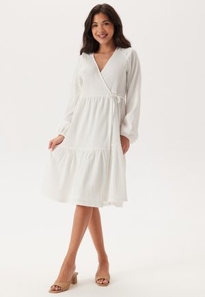 Robe portefeuille blanche en tissu texturé, décolleté en V, longues manches bouffantes et jupe à volants. Elle présente une ceinture en tissu à la taille.