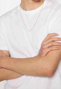 Personne portant un t-shirt blanc uni et un collier en chaîne dorée fine, les bras croisés sur la poitrine.