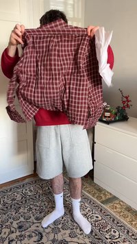 Red and white checkered button-up shirt held in hands, featuring a relaxed fit and long sleeves. Light gray shorts and white socks worn below.