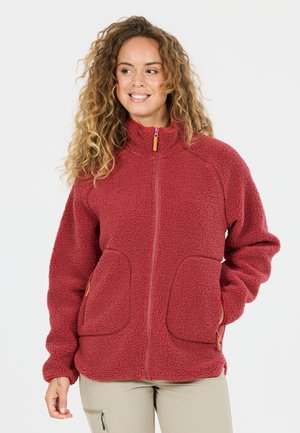 Vrouw met krullend haar draagt een rode fleecejas met rits en twee voorzakken en beige broek, glimlachend en iets naar links kijkend.