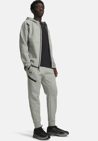 Grauer Kapuzenpullover mit Reißverschluss und seitlichen Taschen, kombiniert mit einem schwarzen T-Shirt und passenden grauen Jogginghosen mit Reißverschlusstaschen. Schwarze Sportschuhe.