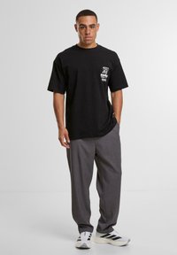 Tricou oversized negru cu logo grafic pe piept, asortat cu pantaloni gri sobru și adidași albi cu dungi negre.
