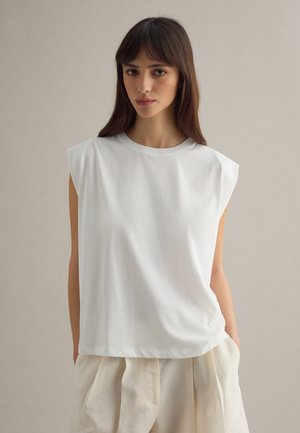 Giovane donna con lunghi capelli scuri, indossa una canottiera bianca e pantaloni beige larghi, in piedi davanti a uno sfondo neutro e semplice.