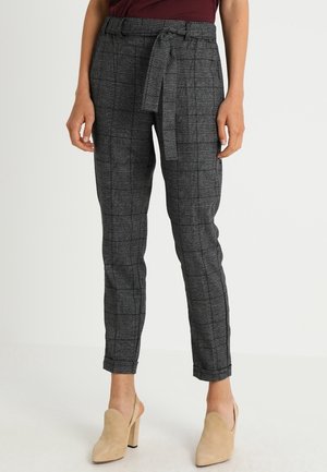 Pantalons à carreaux gris avec une ceinture, coupe ajustée et texture lisse. Portés avec des mules nude, présentant un design à dos ouvert.