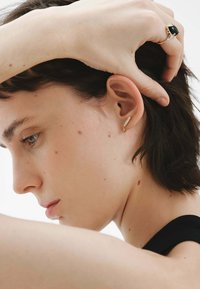 Pendiente de oro parecido a una pluma, colocado en el lóbulo de la oreja. Piel suave con ligeras pecas; cabello corto y oscuro. Fondo neutro.