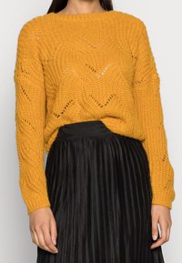 Gelber Strickpullover mit Zickzackmuster und lockerer Passform, kombiniert mit einem schwarzen plissierten Rock. Der Pullover hat lange Ärmel und einen runden Halsausschnitt.