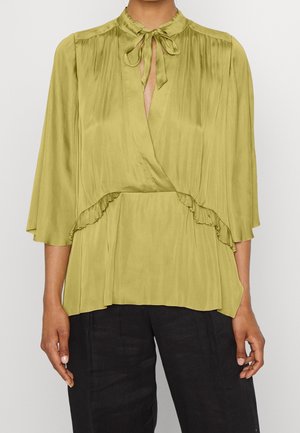 Kvinde iført en lys grøn satinbluse med en bundet sløjfe i halsen, 3/4 ærmer, detaljer med flæser ved taljen, kombineret med sorte bukser.