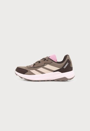 Brązowy i beżowy sneaker Adidas Terrex z różowym językiem, białą podeszwą i trzema beżowymi paskami po boku, pokazany z profilu na białym tle.