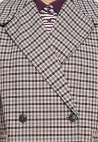 Veste à carreaux croisée avec des boutons noirs, portée sur une chemise rayée bordeaux et blanche à col avec boutons.