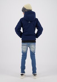 Giacca a vento blu navy con cappuccio e logo triangolare sul retro, abbinata a jeans azzurri e sneaker bianco e nero.