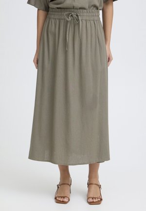 Femme portant une longue jupe vert olive à taille coulissante et des sandales marron à talons ouverts, debout sur un fond blanc uni.