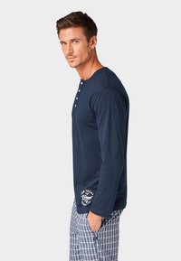 Langarmlige marineblauwe henley shirt, voorzien van een knopenlijst, zacht materiaal en witte logo-details aan de zoom. Gecombineerd met geruite shorts.