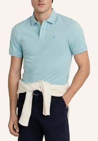 Muž v světle modrém polokošili a tmavých kalhotách s bílým svetrem přehoděným kolem pasu, stojící před jednobarevným pozadím.