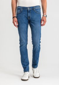 Mężczyzna ubrany w obcisłe niebieskie dżinsy, białe trampki, białą koszulę, srebrny zegarek na prawej ręce i czarną opaskę na nadgarstek na lewej ręce.