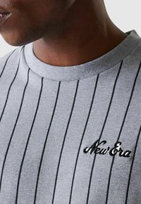 T-shirt gris à manches courtes avec des rayures verticales noires. Dispose d'un col rond et du logo brodé "New Era" sur la poitrine gauche. Texture lisse.